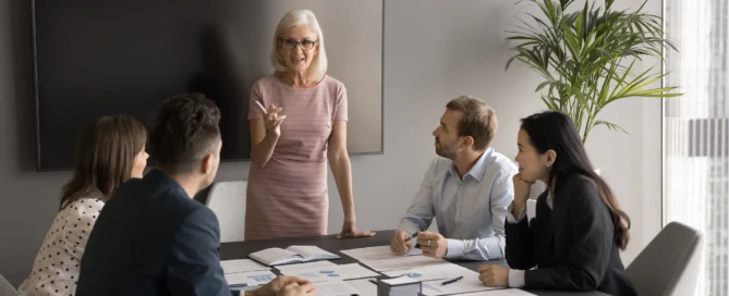 Senior woman in pink dress speaks at a conference table as four colleagues listen, papers spread across the table.